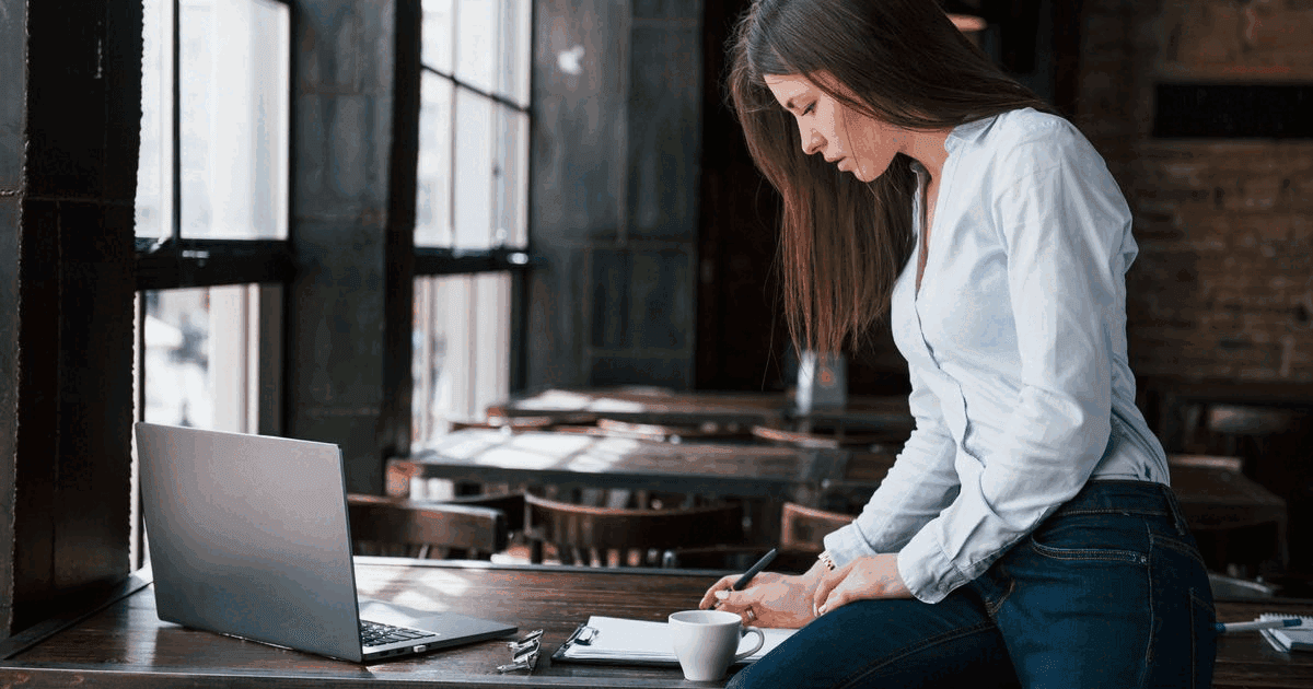 Cafe owner standing at a table in her empty cafe, placing an order on a laptop and noting it down