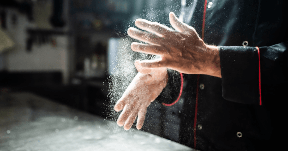 Close-up of a chef's hands clapping flour over a stainless steel kitchen surface
