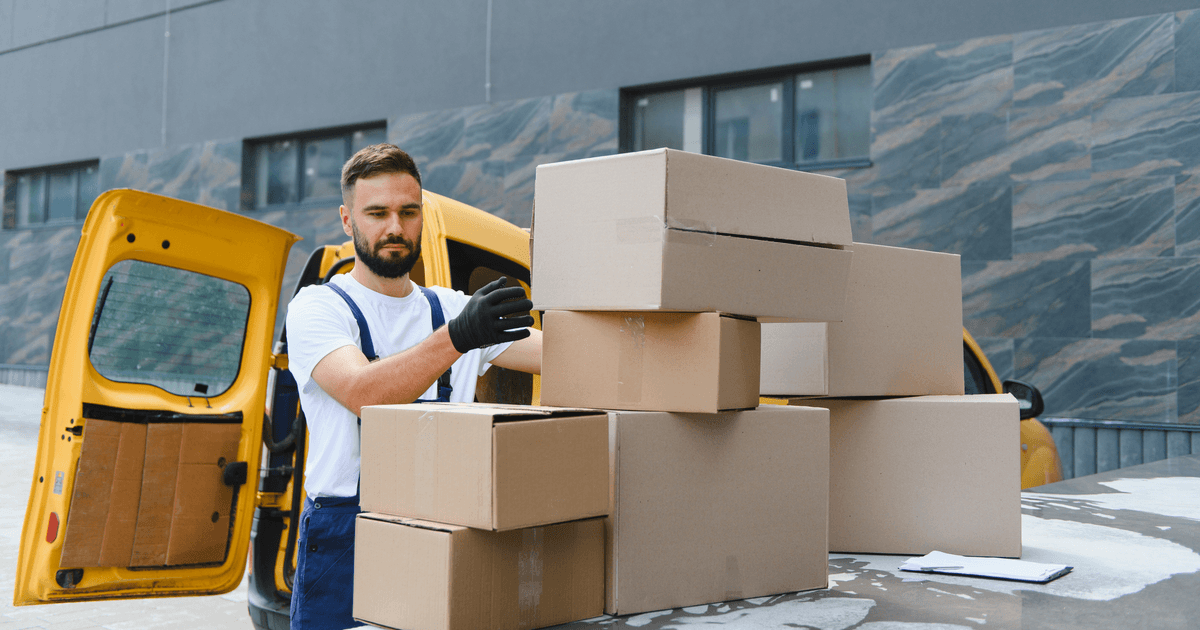 Courier in dungarees loading a stack of cardboard boxes into a yellow delivery van