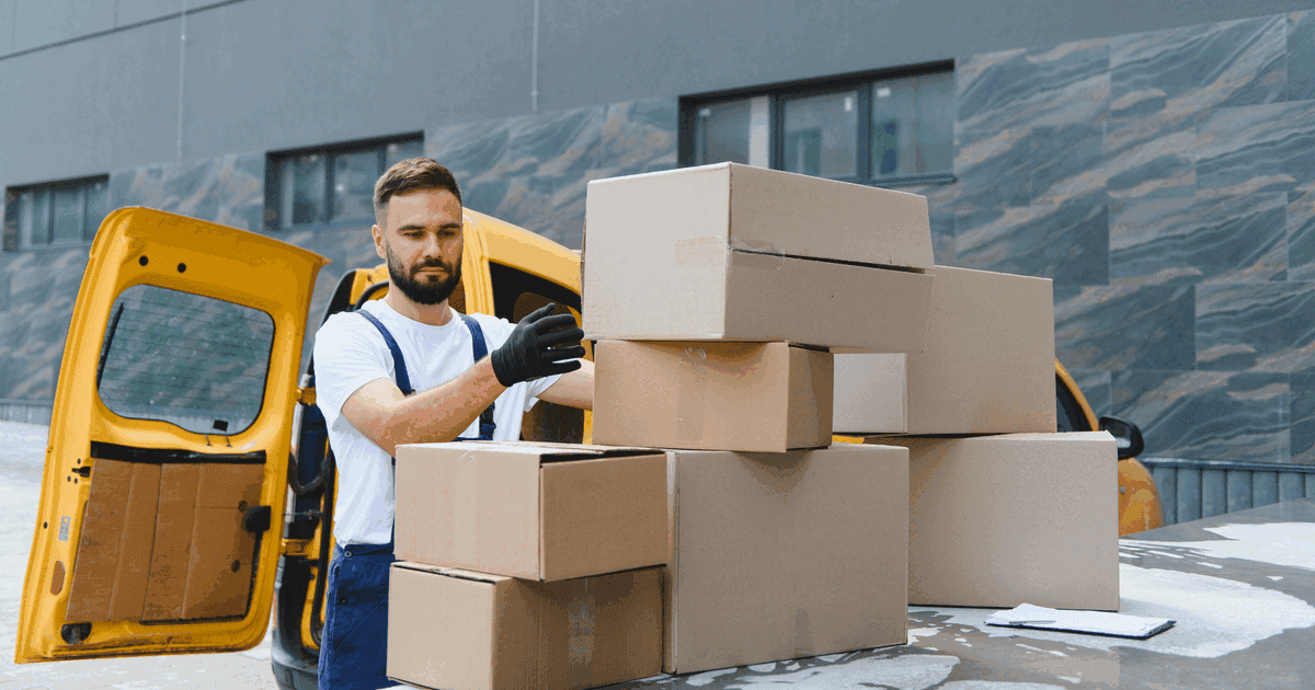 Courier in dungarees loading a stack of cardboard boxes into a yellow delivery van