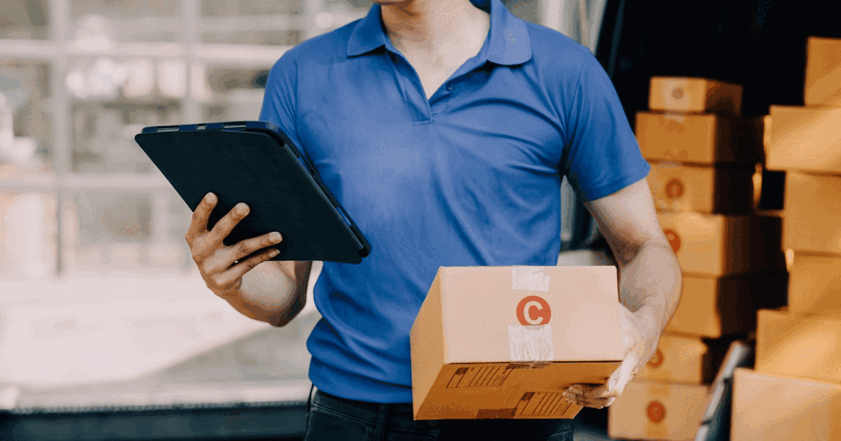 Delivery worker in a blue polo shirt holding a tablet and a cardboard box, with stacks of boxes behind him