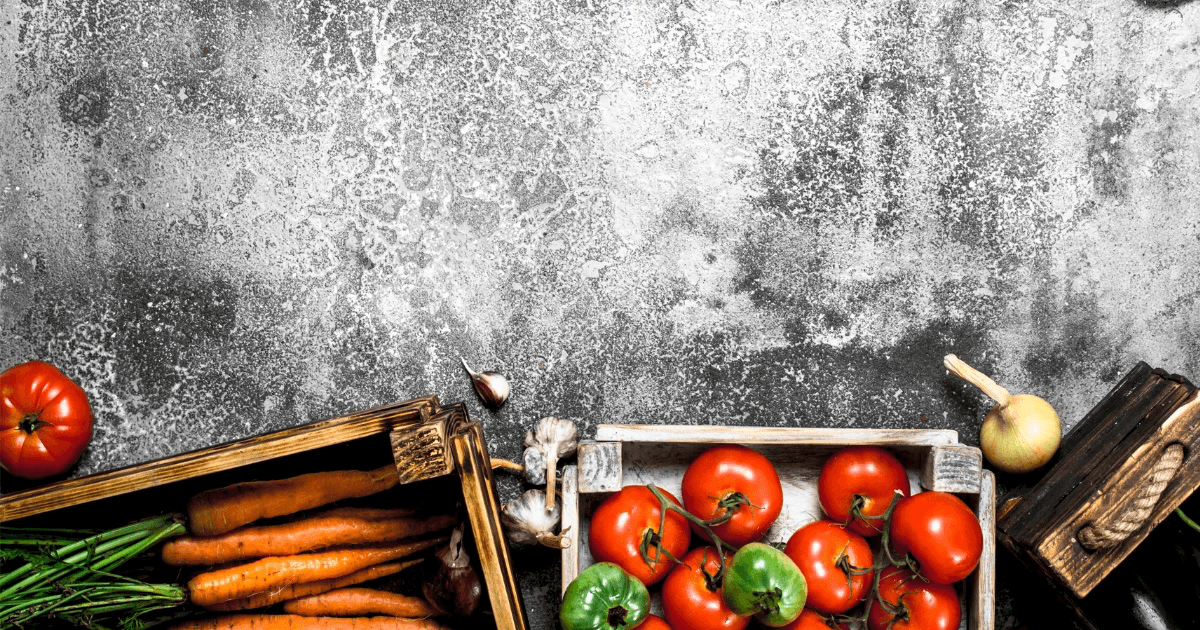 Top-down view of fresh tomatoes, carrots, peppers, onion and garlic in wooden crates on a grey concrete background