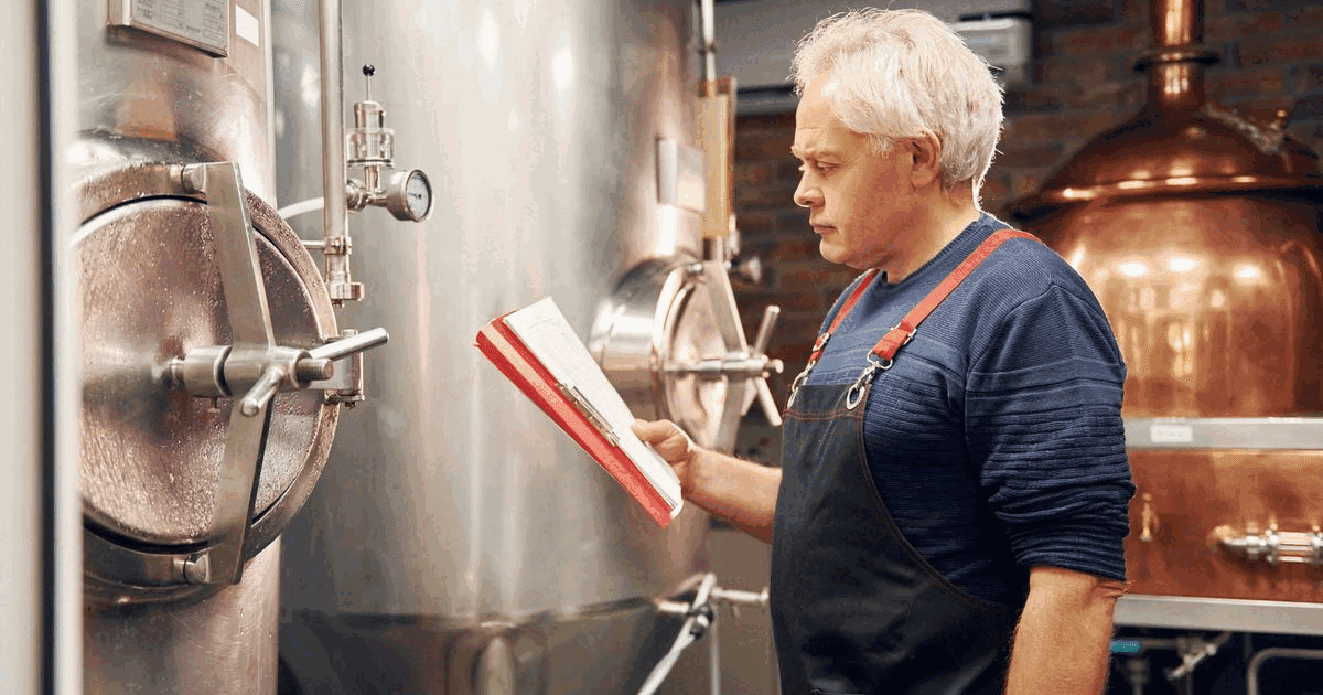 Senior brewer in a brewery storeroom reading an order sheet beside steel fermentation tanks
