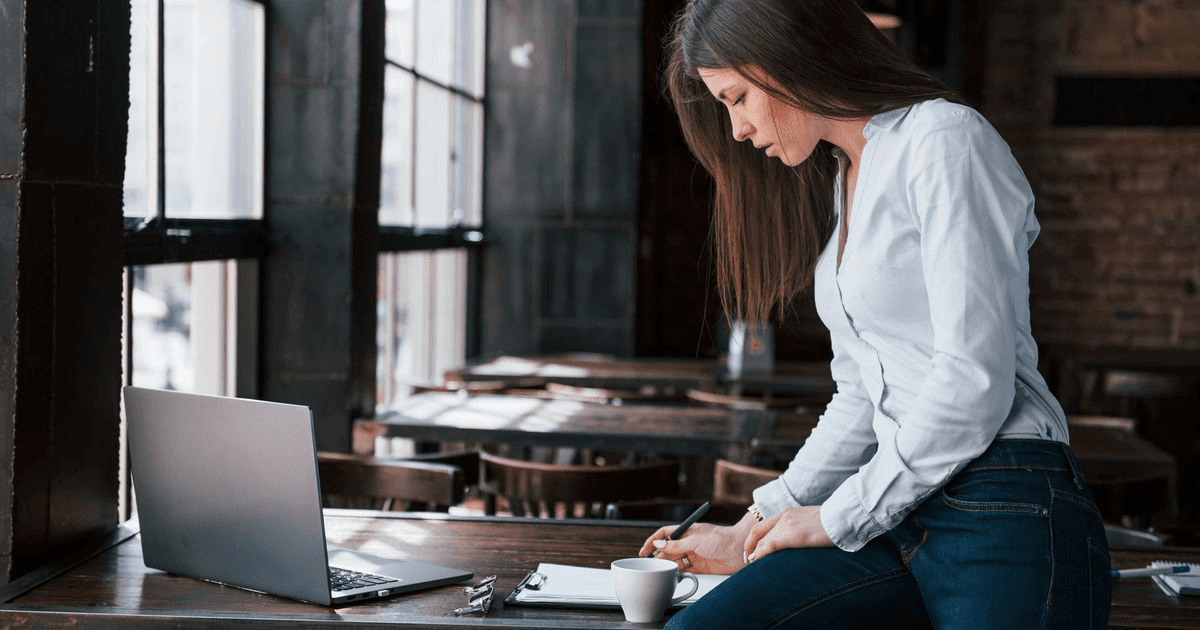 Cafe owner standing at a table in her empty cafe, placing an order on a laptop and noting it down