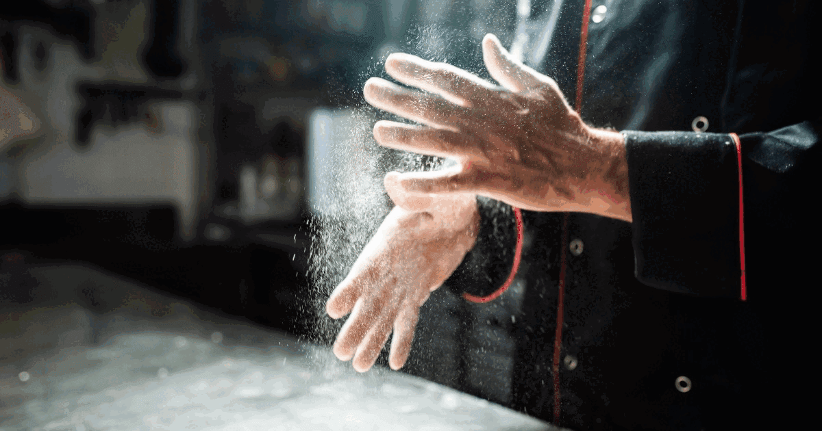 Close-up of a chef's hands clapping flour over a stainless steel kitchen surface