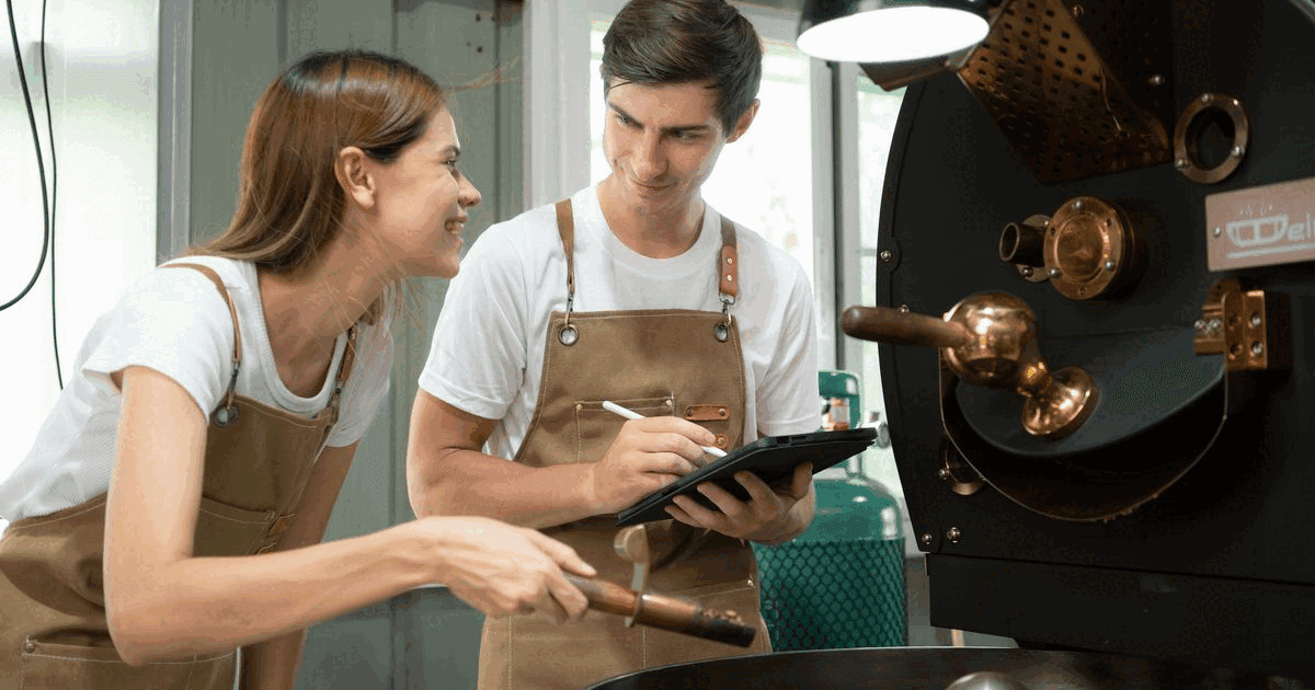 Two coffee roasters in aprons checking an order on a tablet beside a drum roaster