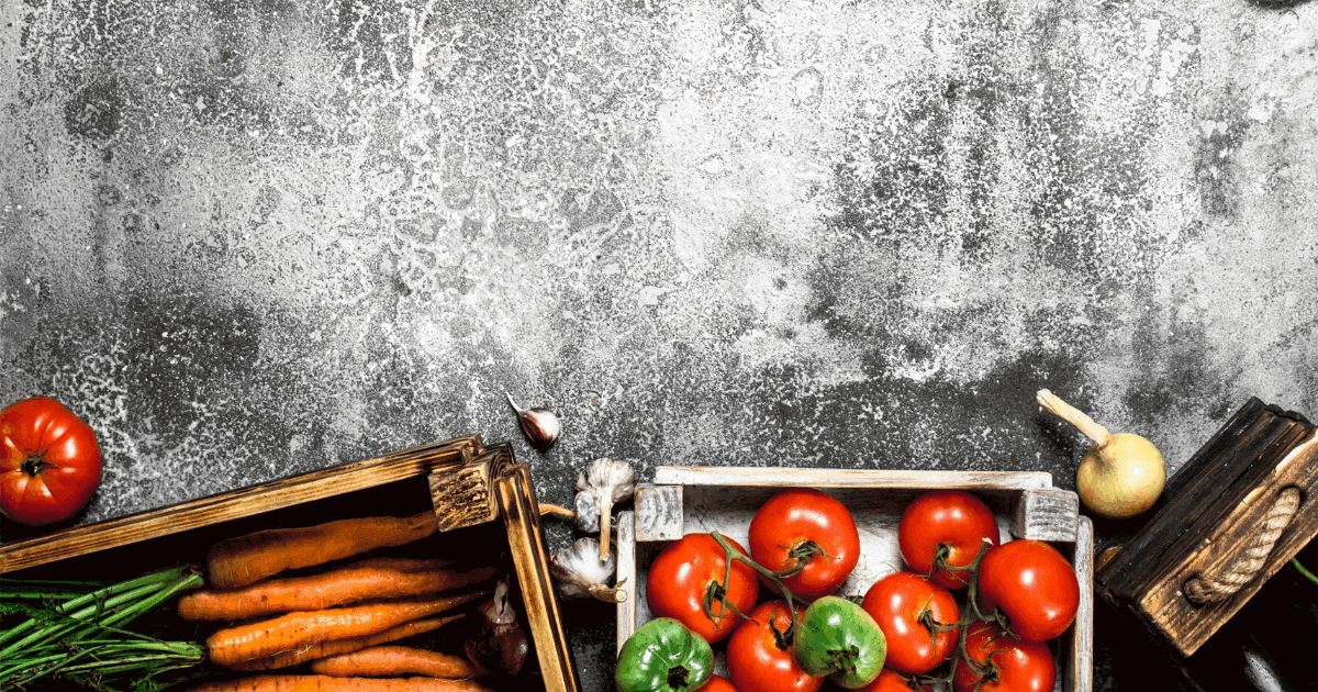 Top-down view of fresh tomatoes, carrots, peppers, onion and garlic in wooden crates on a grey concrete background