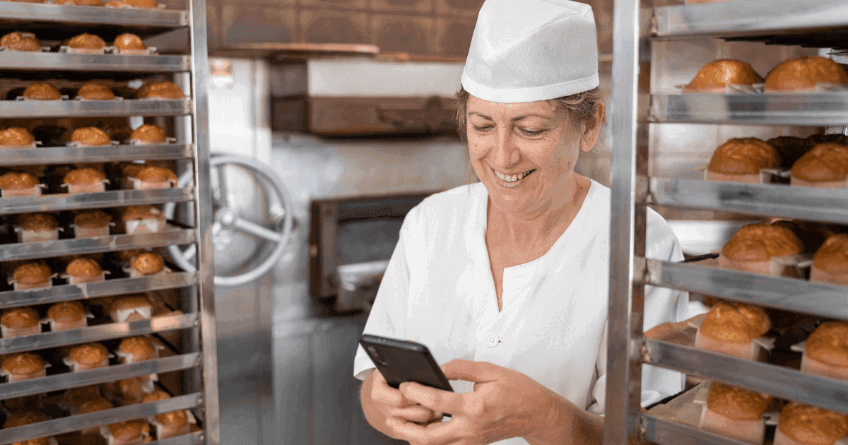 Baker woman smiling and texting with her phone surrounded by fresh baked cupcakes